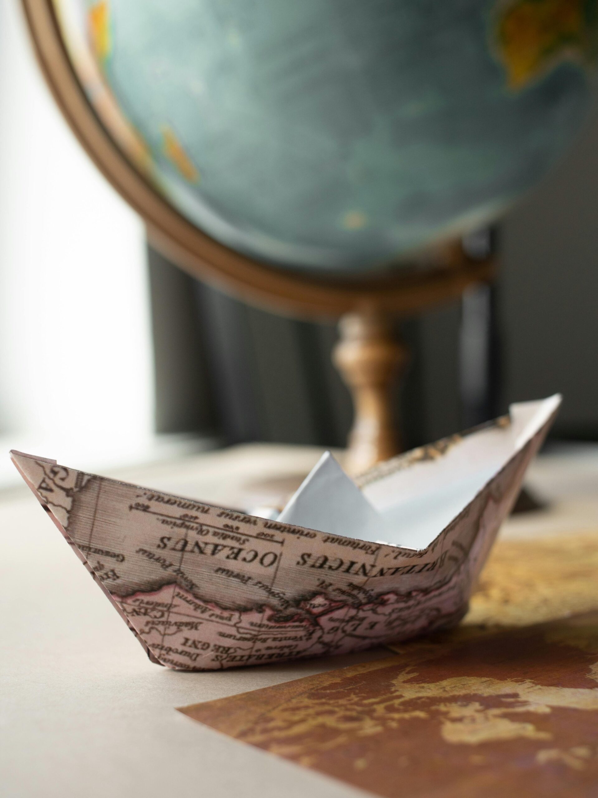 Paper boat made of world map placed on brown wooden table near round globe with continents in light room at home