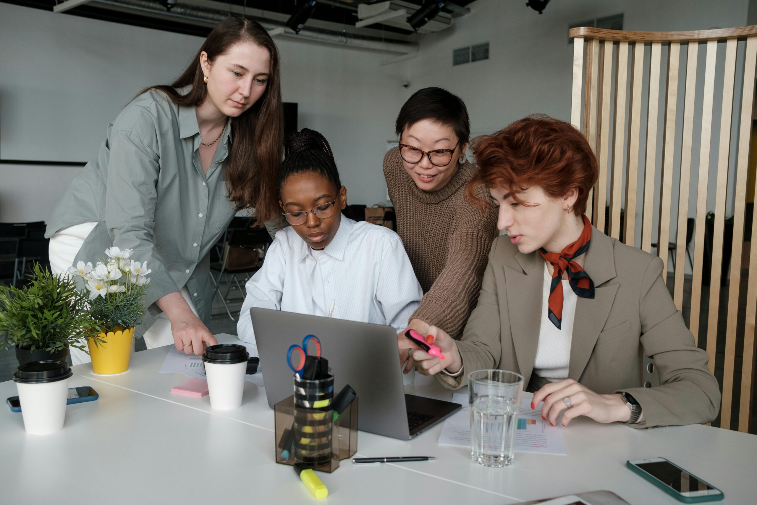A diverse group of women engaged in a business meeting around a laptop in a modern office setting.